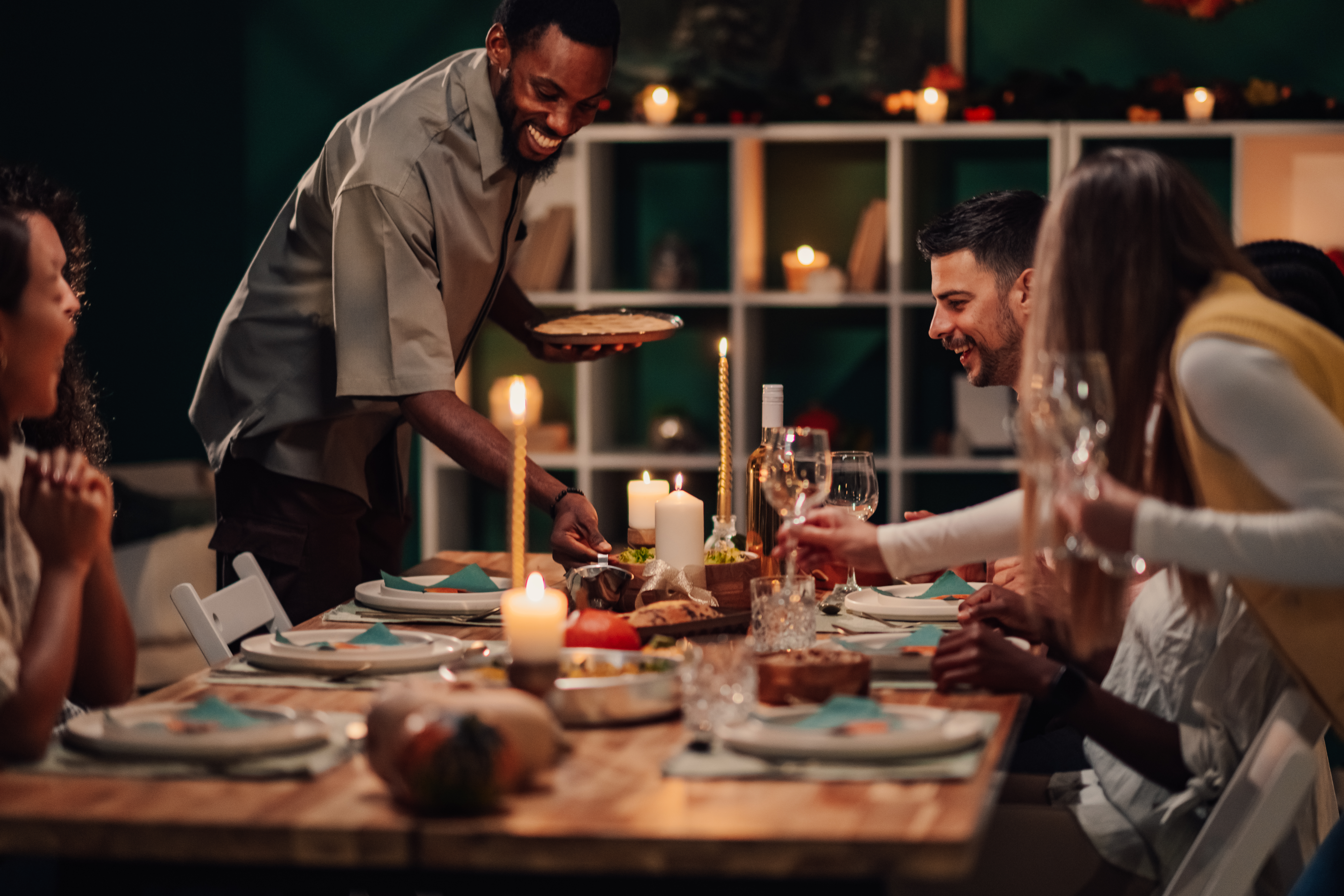Happy friends enjoying festive dinner: host serving food at candlelit table